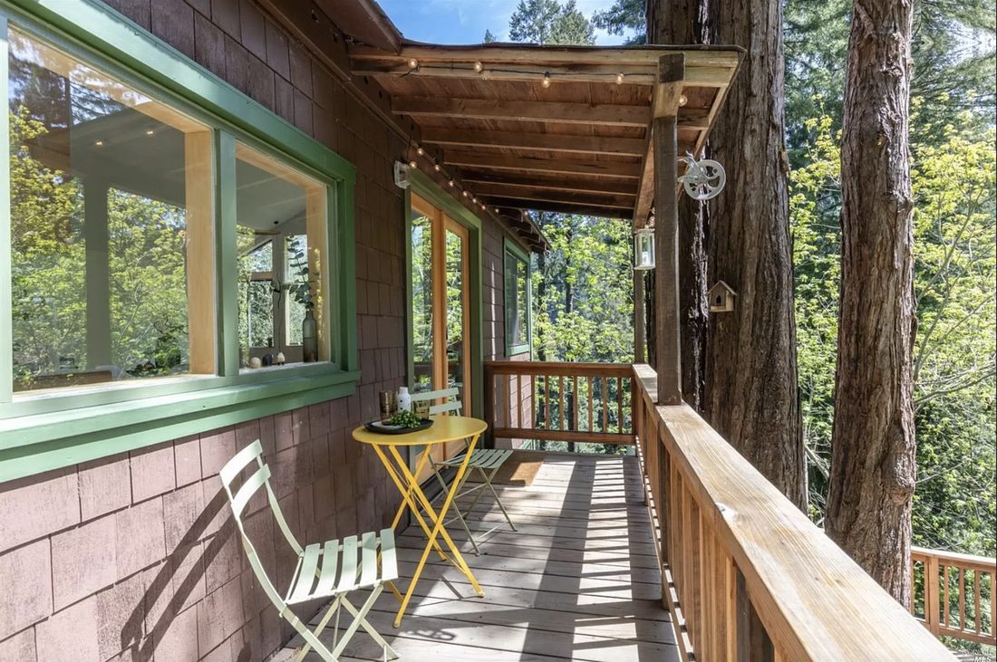A narrow balcony with a yellow bistro set and string lights, running along the green-trimmed shingle wall beside a redwood.