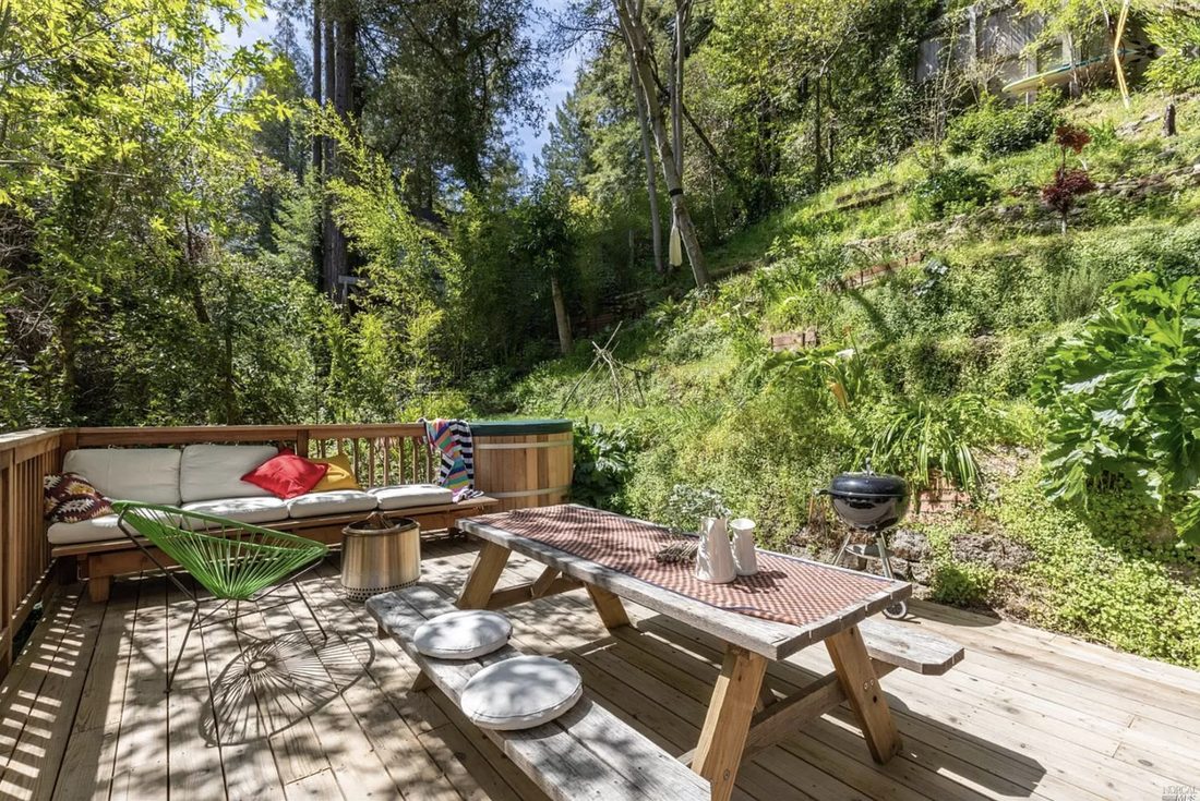 A picnic table and outdoor sofa on a forest deck with a kettle grill nearby.