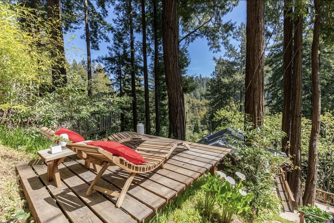 Two wooden lounge chairs with red cushions on a sun-dappled deck looking out over a redwood valley.