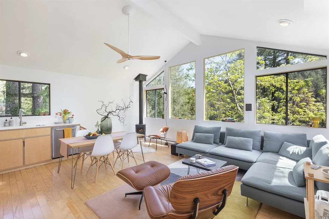 Open-plan great room with a vaulted ceiling, Eames lounge chair, grey sectional, and a wall of windows looking into sunlit trees.