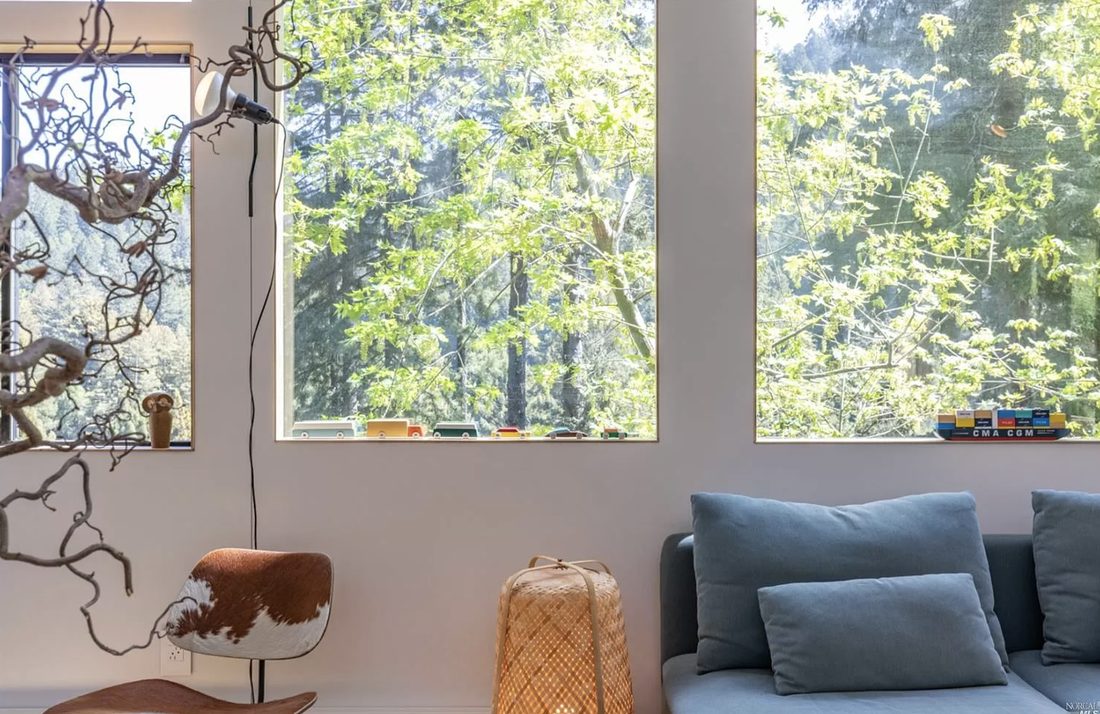 A sunlit corner of the living room with a cowhide chair, a sculptural branch, and tall windows over a blue sofa.