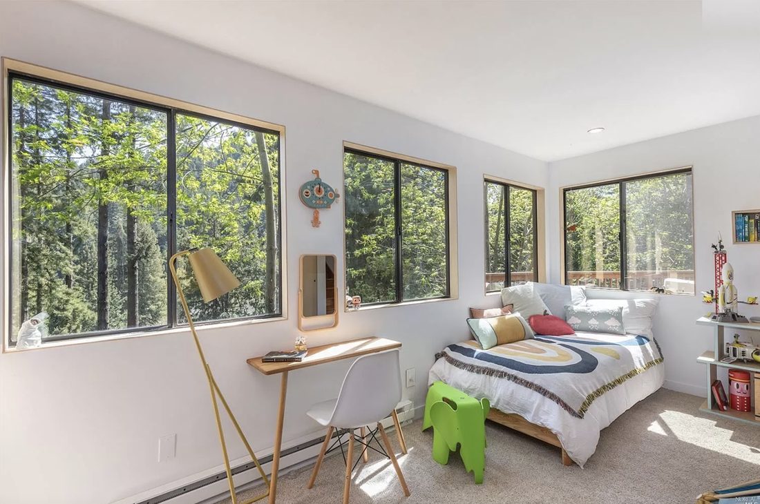 The main bedroom, bright and simple, wrapped on two sides by large windows full of green leaves.