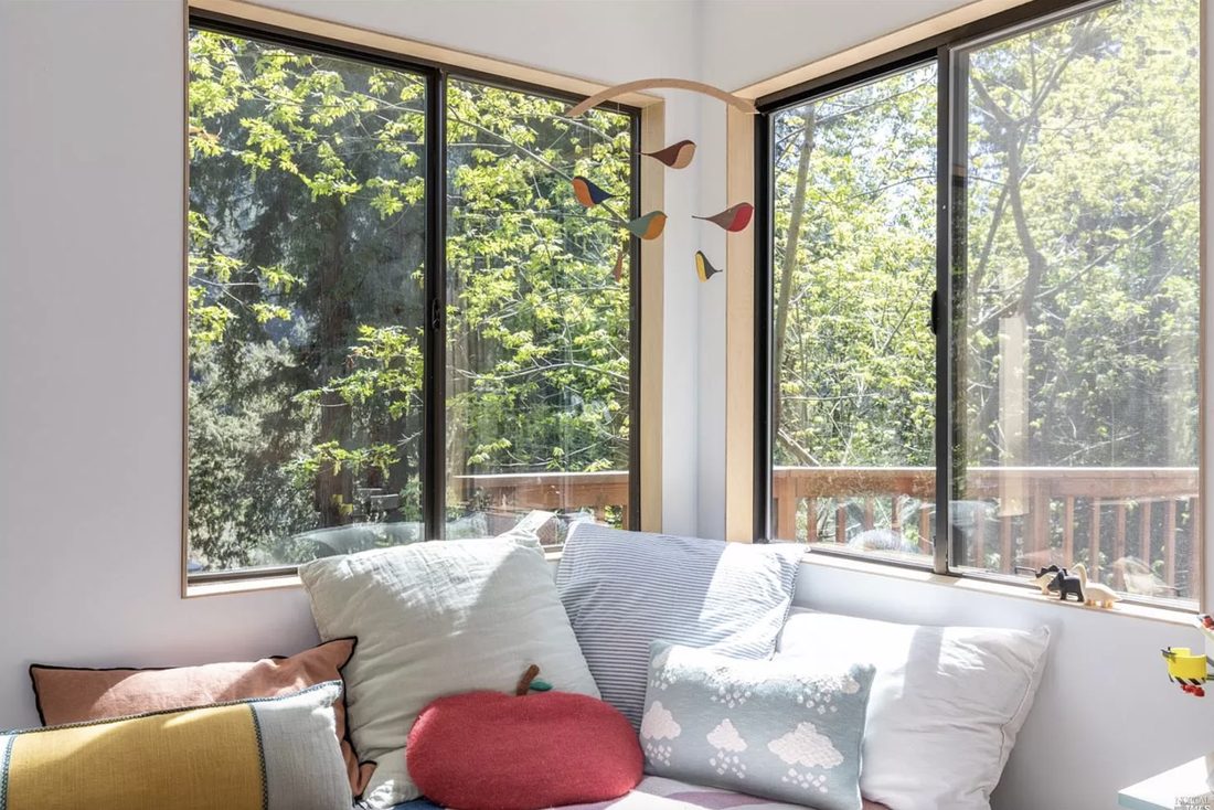 A cushioned window seat layered with linen pillows beneath a corner of windows.